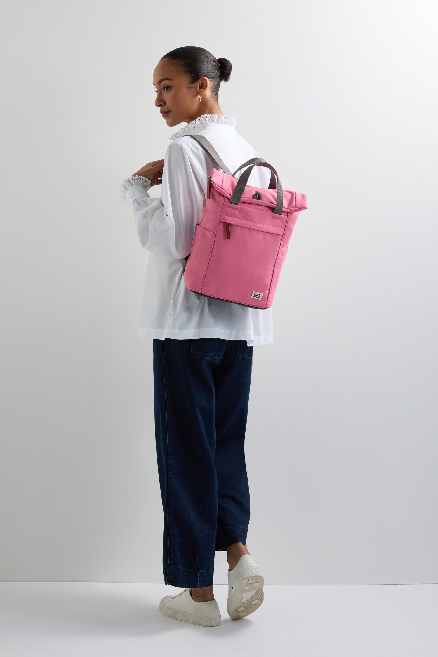 A person in a white blouse and dark pants, with hair tied back, stands against a plain background carrying a pink Recycled Canvas Small Finchley Bag.