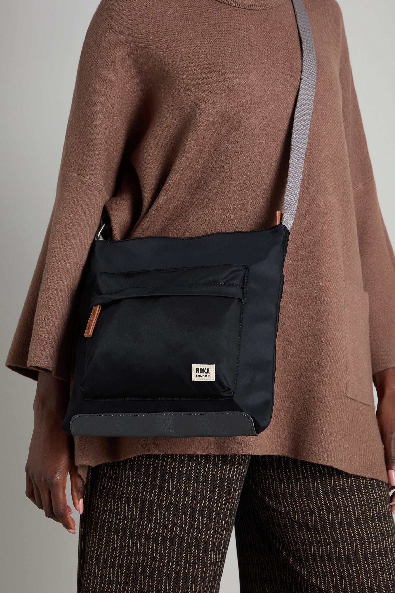 A person dressed in a brown sweater and patterned pants carries the Recycled Nylon Kennington Crossbody Bag in sleek black, which is crafted with a grey strap and pocket detailing.