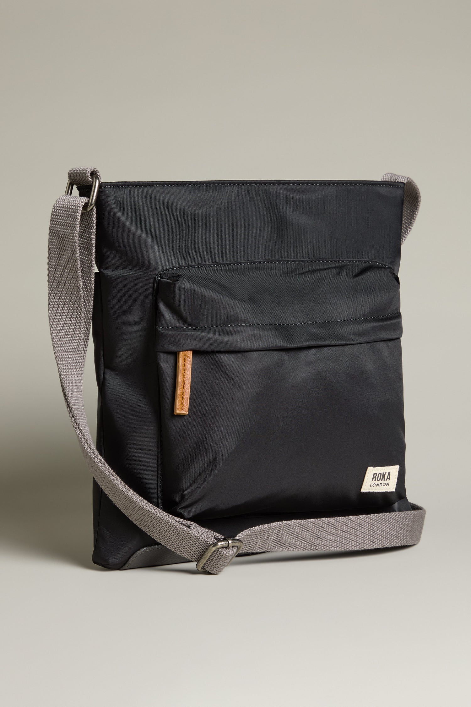 The Recycled Nylon Kennington Crossbody Bag is a sleek black accessory made from water-resistant fabric, featuring a gray adjustable strap and front pocket, set against a neutral background.