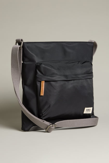 The Recycled Nylon Kennington Crossbody Bag is a sleek black accessory made from water-resistant fabric, featuring a gray adjustable strap and front pocket, set against a neutral background.