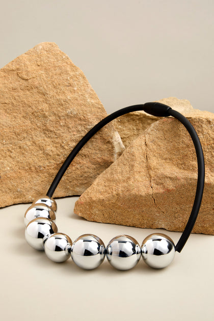 The 7 Spheres Necklace showcases contemporary elegance with its prominent silver spheres delicately positioned on a sleek black band, set against geometric tan stones.