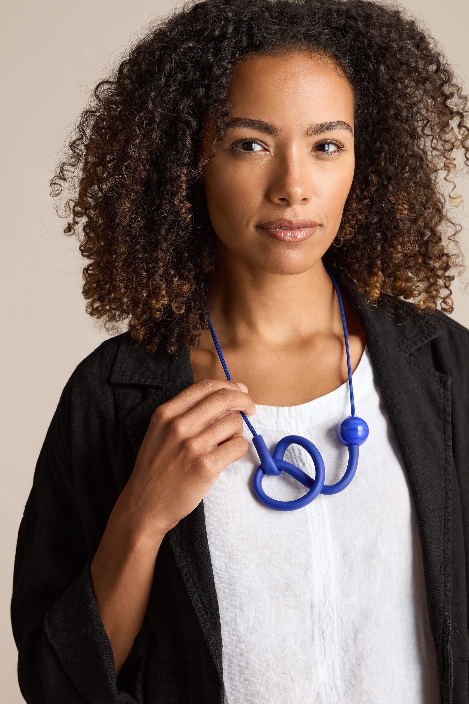 A person with curly hair wearing a white shirt and black jacket, holding the earpiece of a blue stethoscope that hangs around their neck, accessorized with a Murano Glass Knot Necklace.