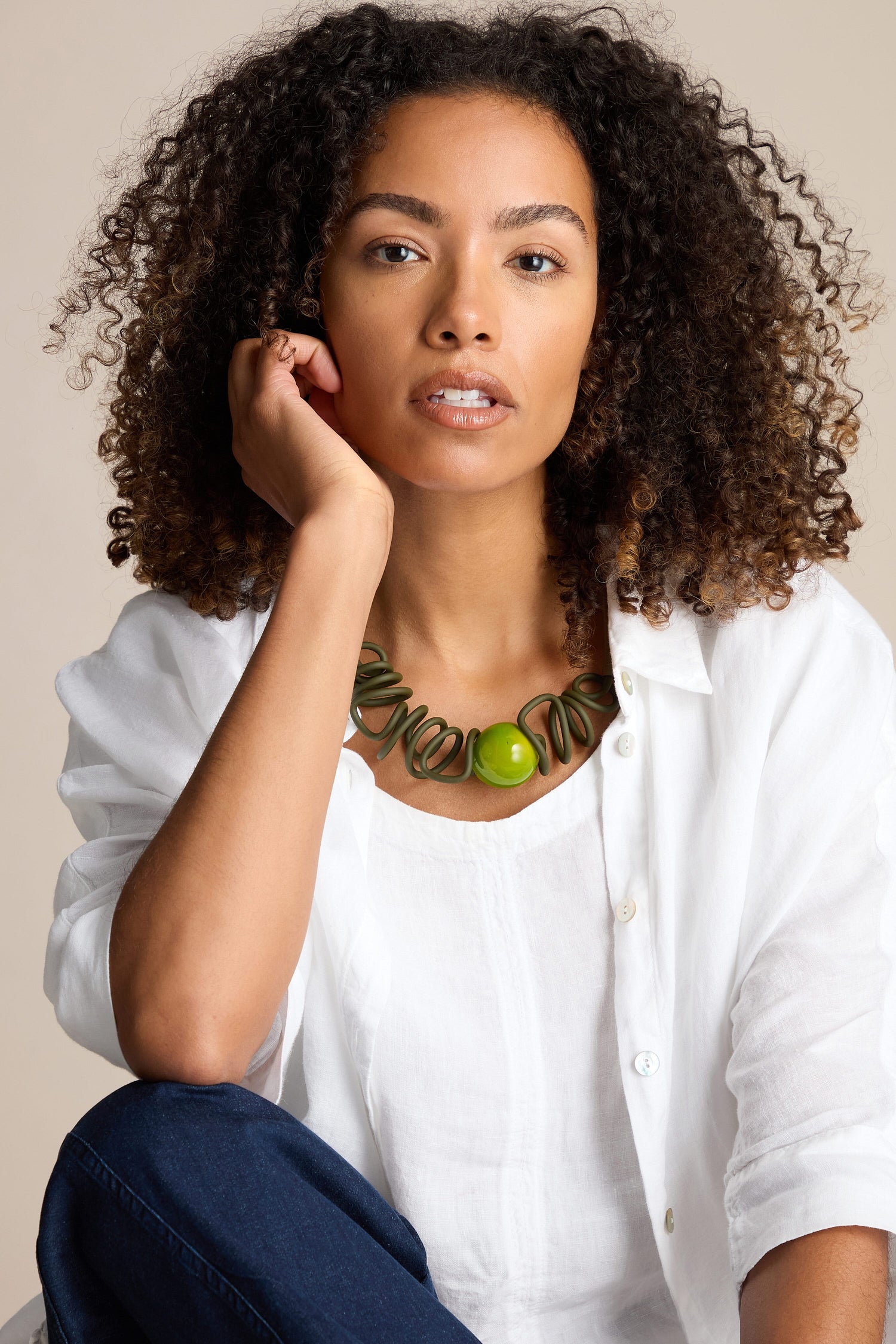 A person with curly hair wearing a white shirt, blue jeans, and a green Murano Sketch Necklace by Samuel Coraux, sitting with one hand touching their face and a neutral expression.