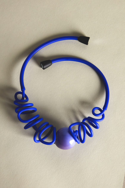 A blue cable with a large purple bead and black end caps, resembling the dramatic color of a Murano Sketch Necklace by Samuel Coraux, elegantly lies coiled on a light beige surface.