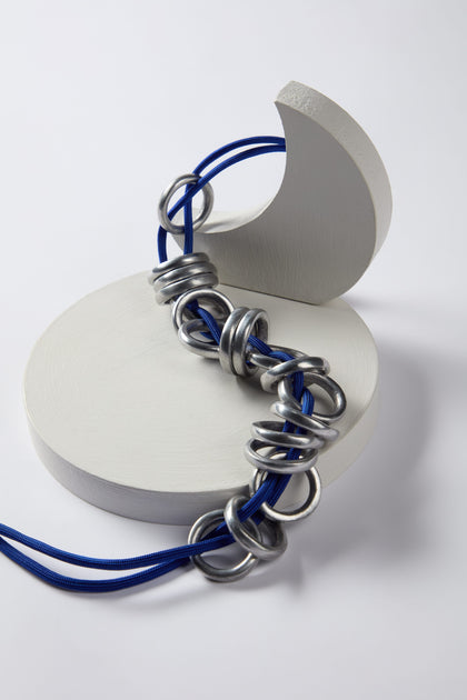 The Metal Rings Necklace by Samuel Coraux features a sculptural design, intricately combining a silver ring chain with a blue cord, artfully draped over white, curved geometric shapes on a white background.