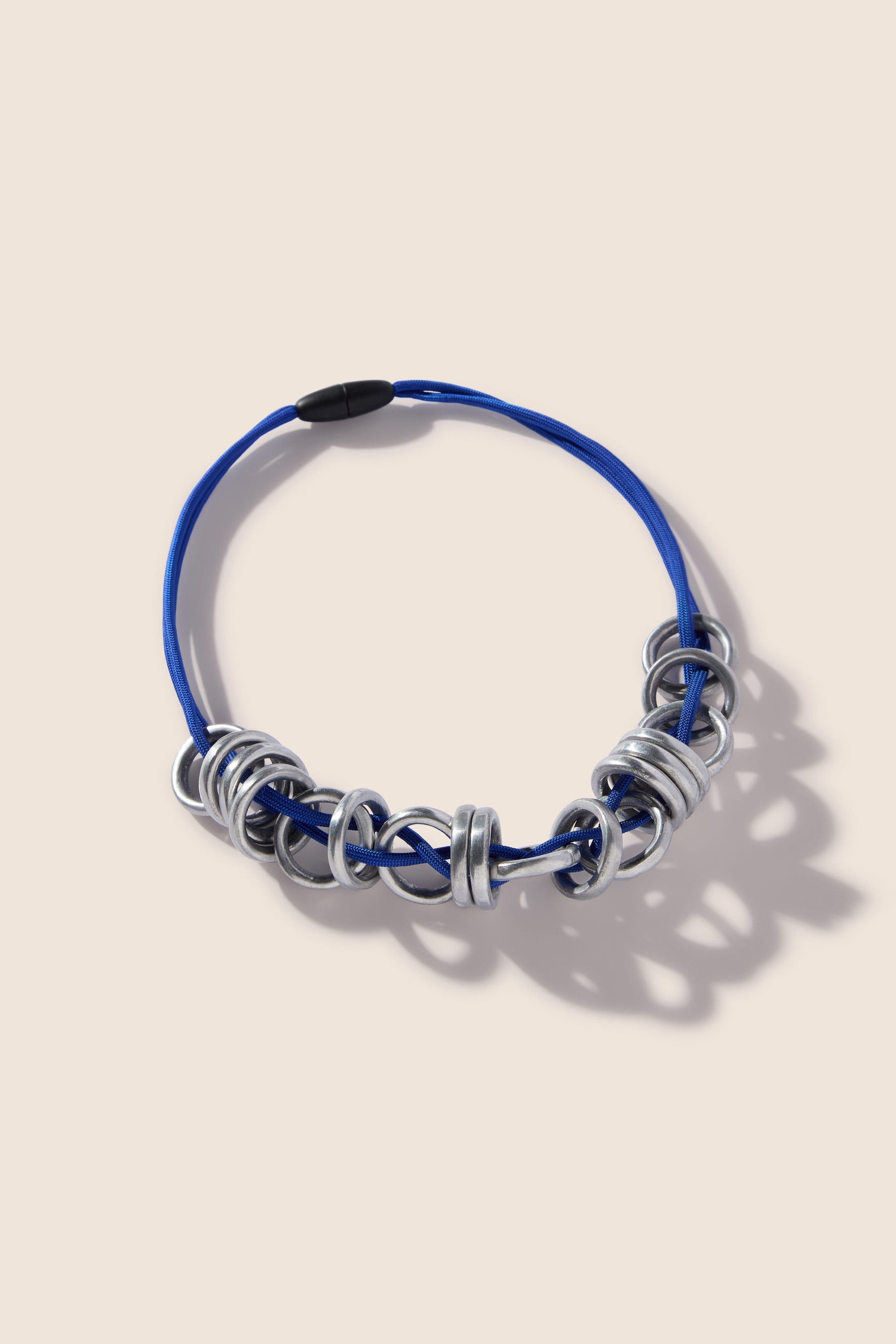 A blue Metal Rings Necklace, featuring multiple silver rings and a black clasp, reminiscent of a Samuel Coraux sculptural accessory, laid flat on a neutral background.