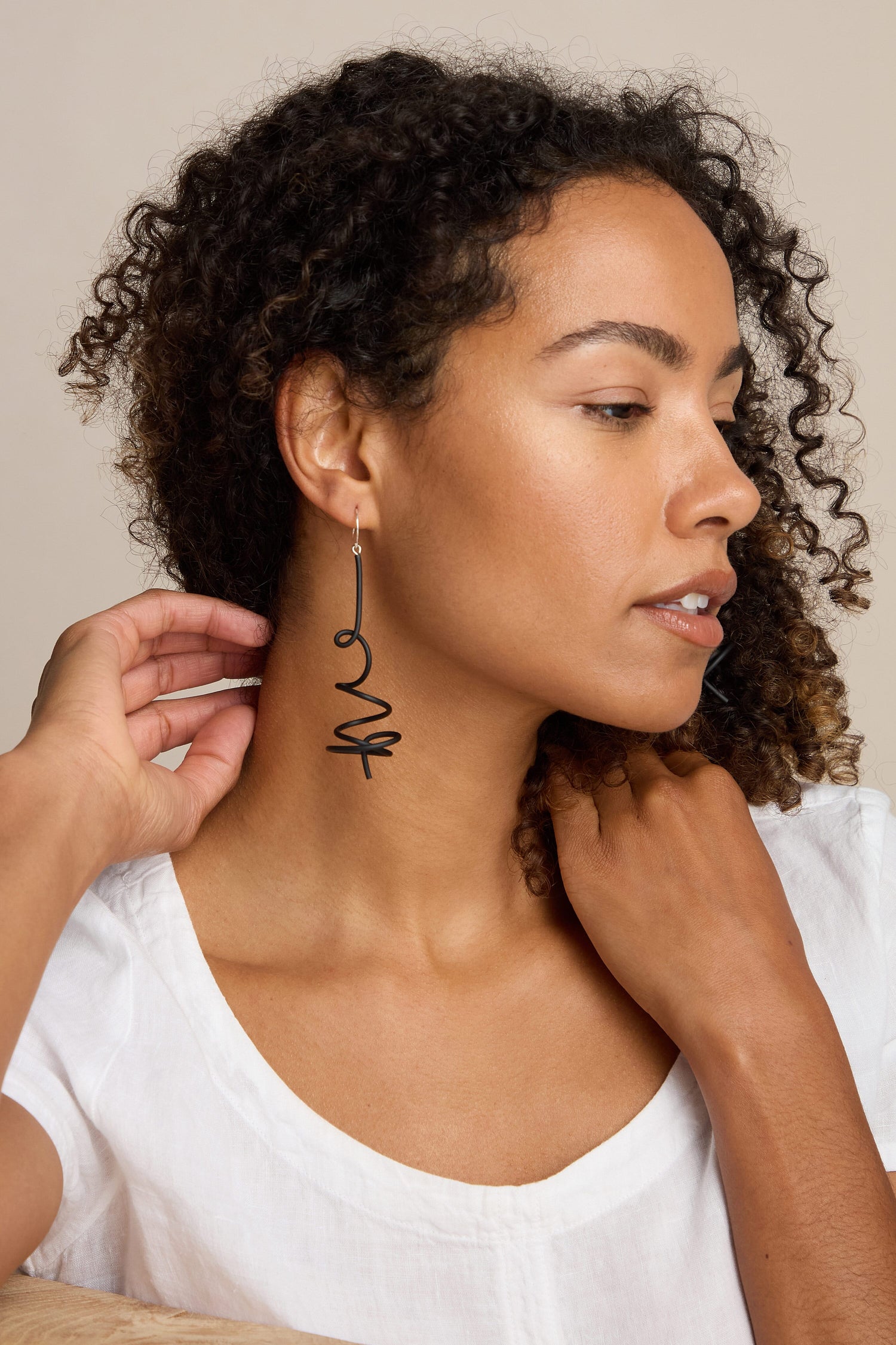 A woman with curly hair is wearing a white shirt and long, playful Squiggle Earrings by Samuel Coraux. She is touching her neck with one hand and looking to the side.