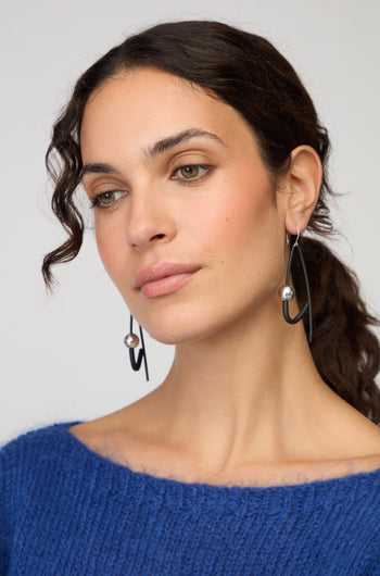 Woman with wavy dark hair wearing a blue sweater and Silver Sphere Earrings by Samuel Coraux—large silver and black drop earrings—looking slightly to the side with a neutral expression.