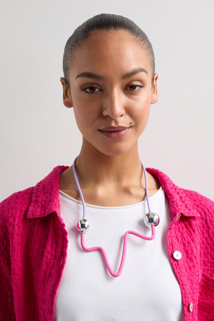 Wearing a vibrant pink jacket and white top, a person sports round earbuds linked by a flexible pink wire around their neck against a plain white background. The look parallels the colorful style of the Silver Sphere Colour Pop Necklace, creating an effortless statement piece.