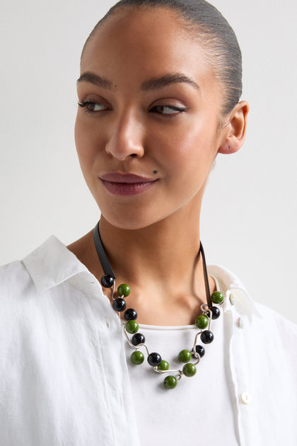 A person in a white shirt showcases the Woven Spheres Necklace by Samuel Coraux, featuring alternating green and black beads that form a sculptural design.