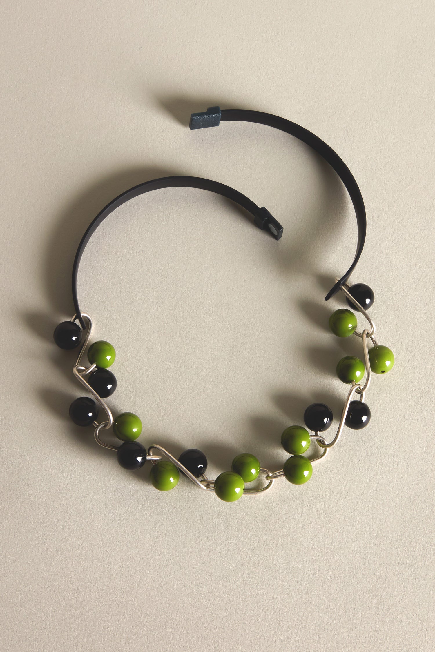 A black headband features alternating black and green beads in a sculptural Samuel Coraux style, joined by a silver wavy wire, displayed on beige. Inspired by the Woven Spheres Necklace design.