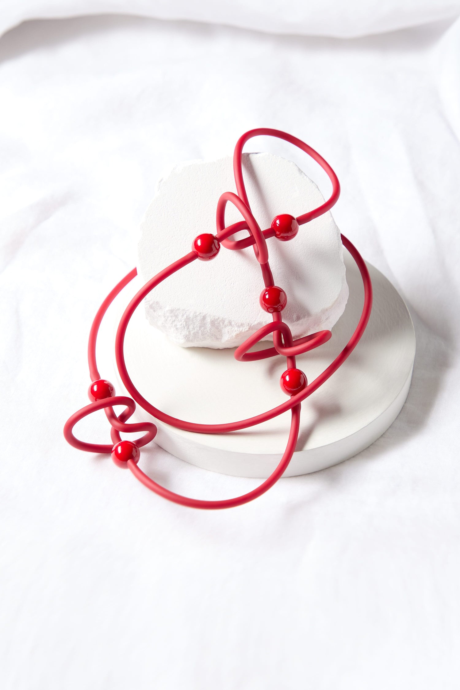 The Trio Squiggle Necklace, inspired by Samuel Coraux, features red looping curves and beads set against a white circular base, capturing the dynamic essence of abstract design.