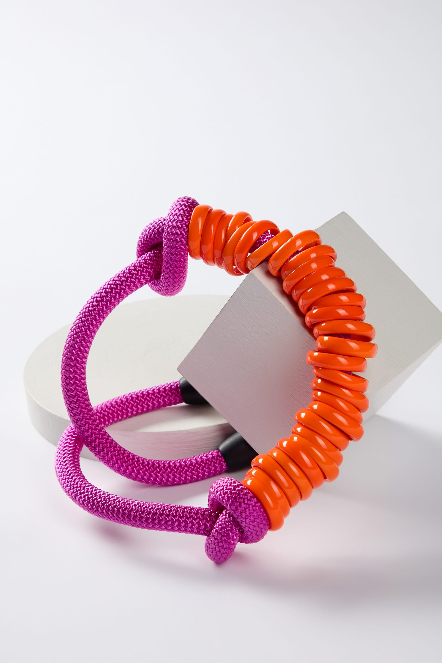 A Samuel Coraux creation, a purple braided cord and orange disc bracelet displayed with geometric blocks against a white background, alongside a Murano Glass Rings Necklace in fuchsia pink.