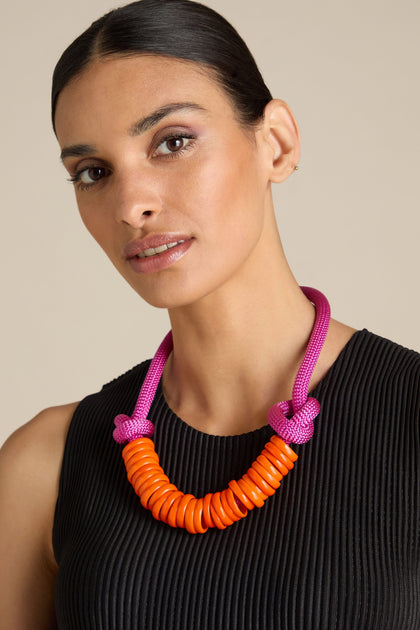 Person wearing a black sleeveless top and a colorful Murano Glass Rings Necklace by Samuel Coraux with orange beads and a pink cord. This ensemble, also featuring vibrant Murano glass rings, adds an extra touch of elegance.