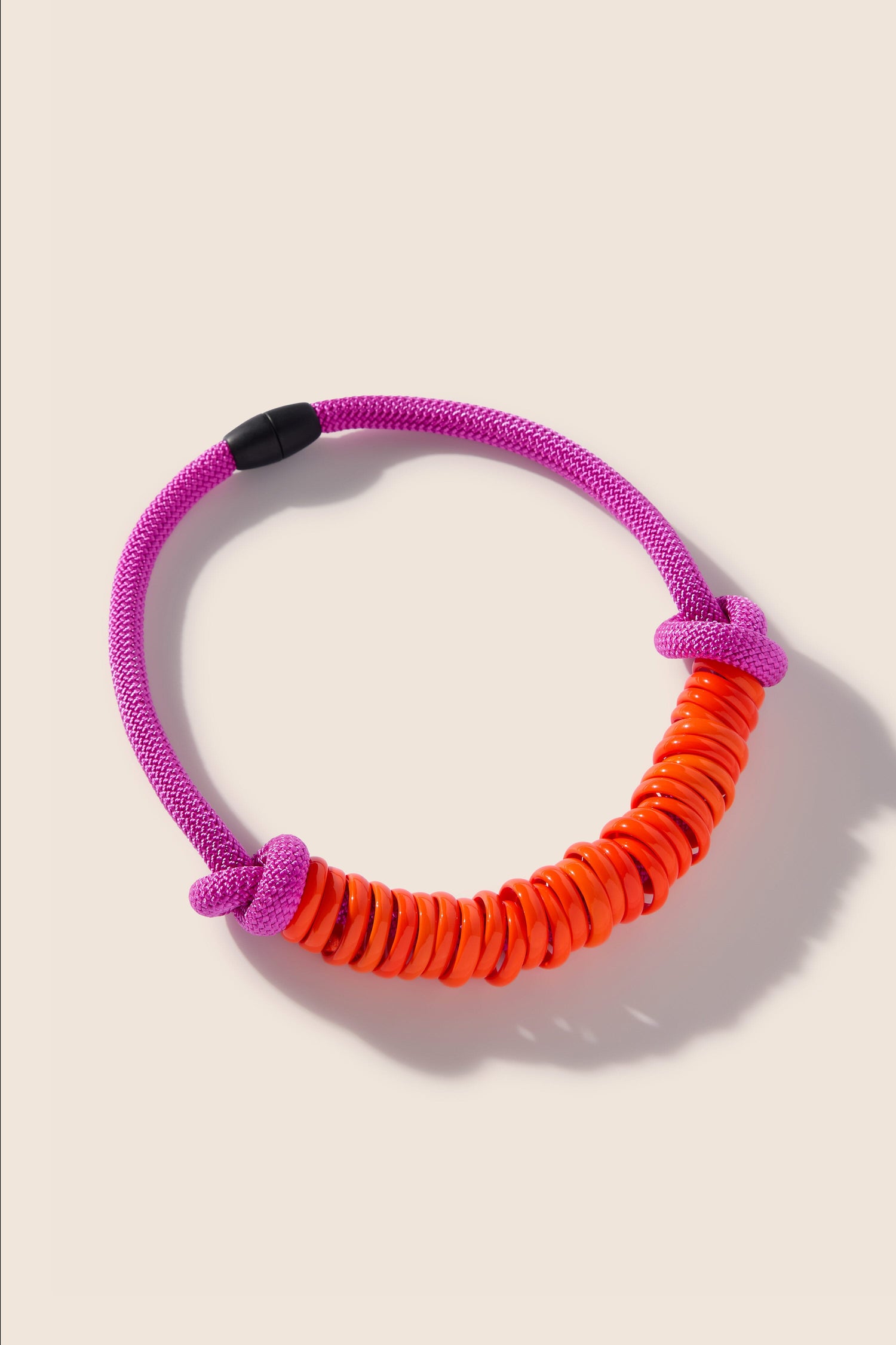 A beige background showcases a Murano Glass Rings Necklace, featuring a complementary purple cord bracelet adorned with orange circular beads and a black clasp, highlighted by hints of fuchsia pink throughout.