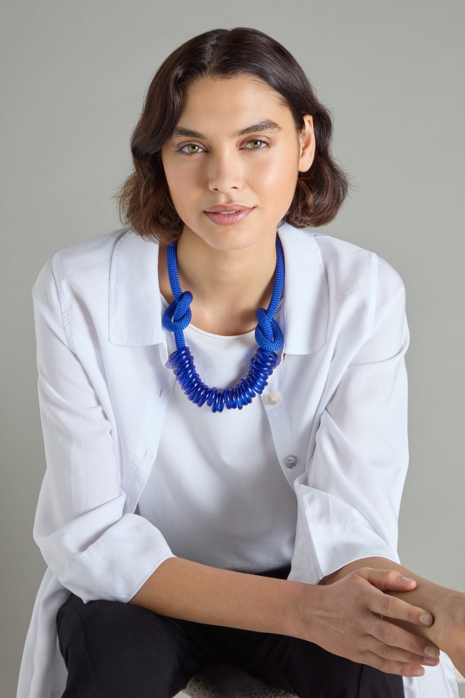 A person wearing a white shirt and a striking blue Murano Glass Rings Necklace by Samuel Coraux sits gracefully, gazing at the camera against a plain backdrop.