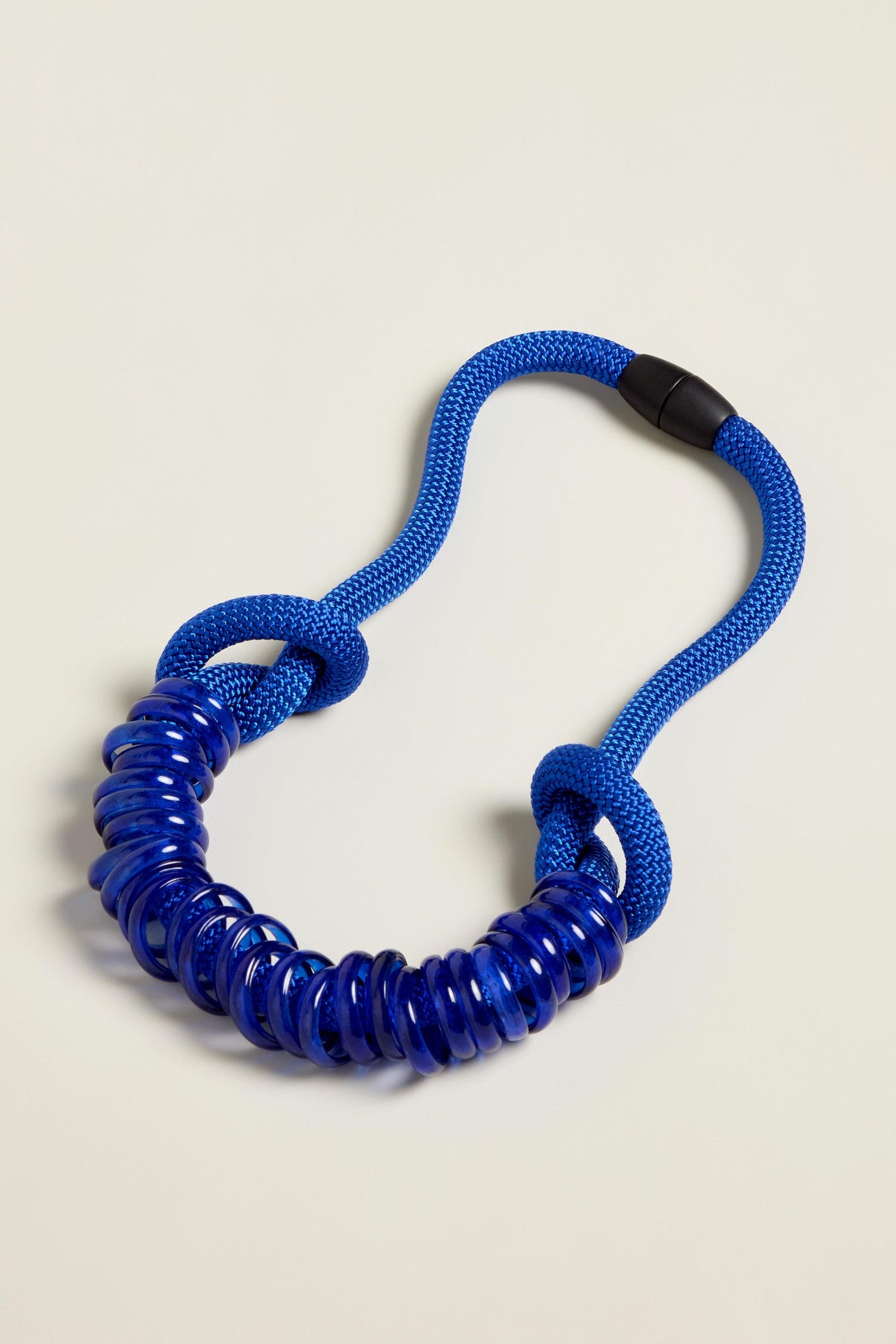 The Murano Glass Rings Necklace, with its sleek coiled blue rubber design, intertwines seamlessly with a textured blue rope on a light surface, evoking the styling of Samuel Coraux's contemporary necklaces.