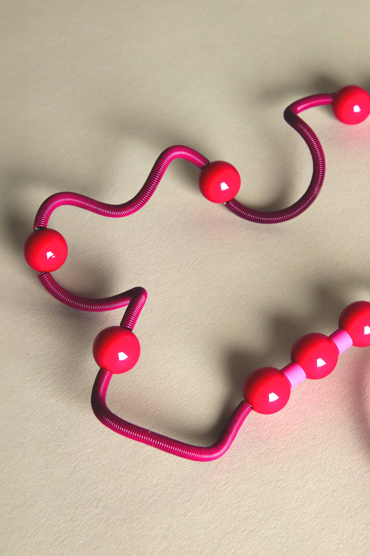 On a beige surface rests the Fuchsia Spiral Glass Spheres Necklace, featuring a curved pink wire adorned with eight red beads that reflect a French jewellery designer's eccentric flair.