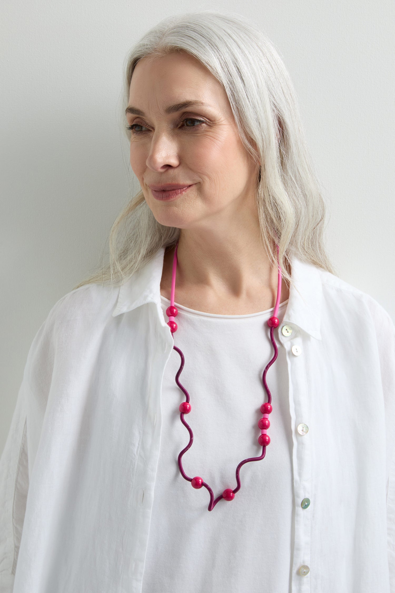 A woman with long gray hair wears a white shirt and a Fuchsia Spiral Glass Spheres Necklace created by an eccentric French jewellery designer, standing against a plain white background.