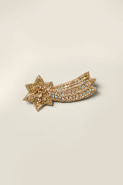 The Shooting Star Brooch is a celestial charm, featuring a gold brooch in the shape of a shooting star with shimmering embedded gemstones on a sleek backdrop—a truly sparkling accessory.