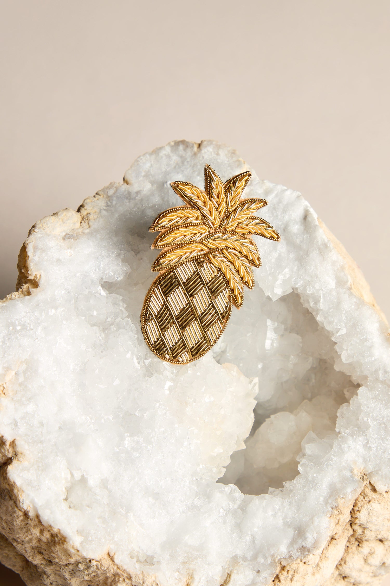 The Pineapple Brooch, showcased on a white geode rock against a beige background, features intricate golden beads and offers a subtle tropical flair.