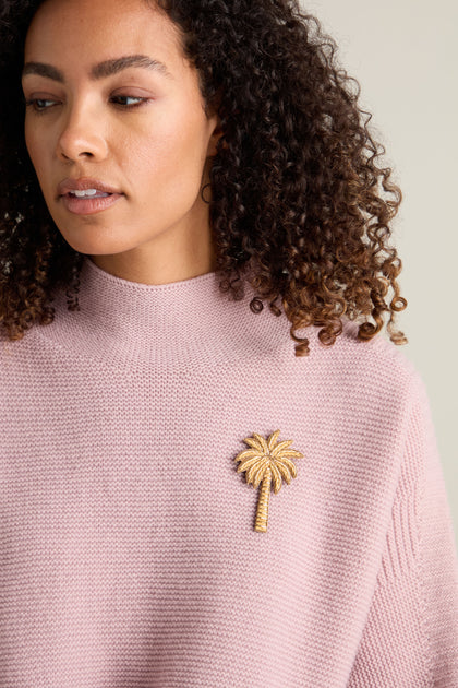 A woman with curly hair dons a pink sweater, accentuated by the glamorous Golden Palm Brooch.