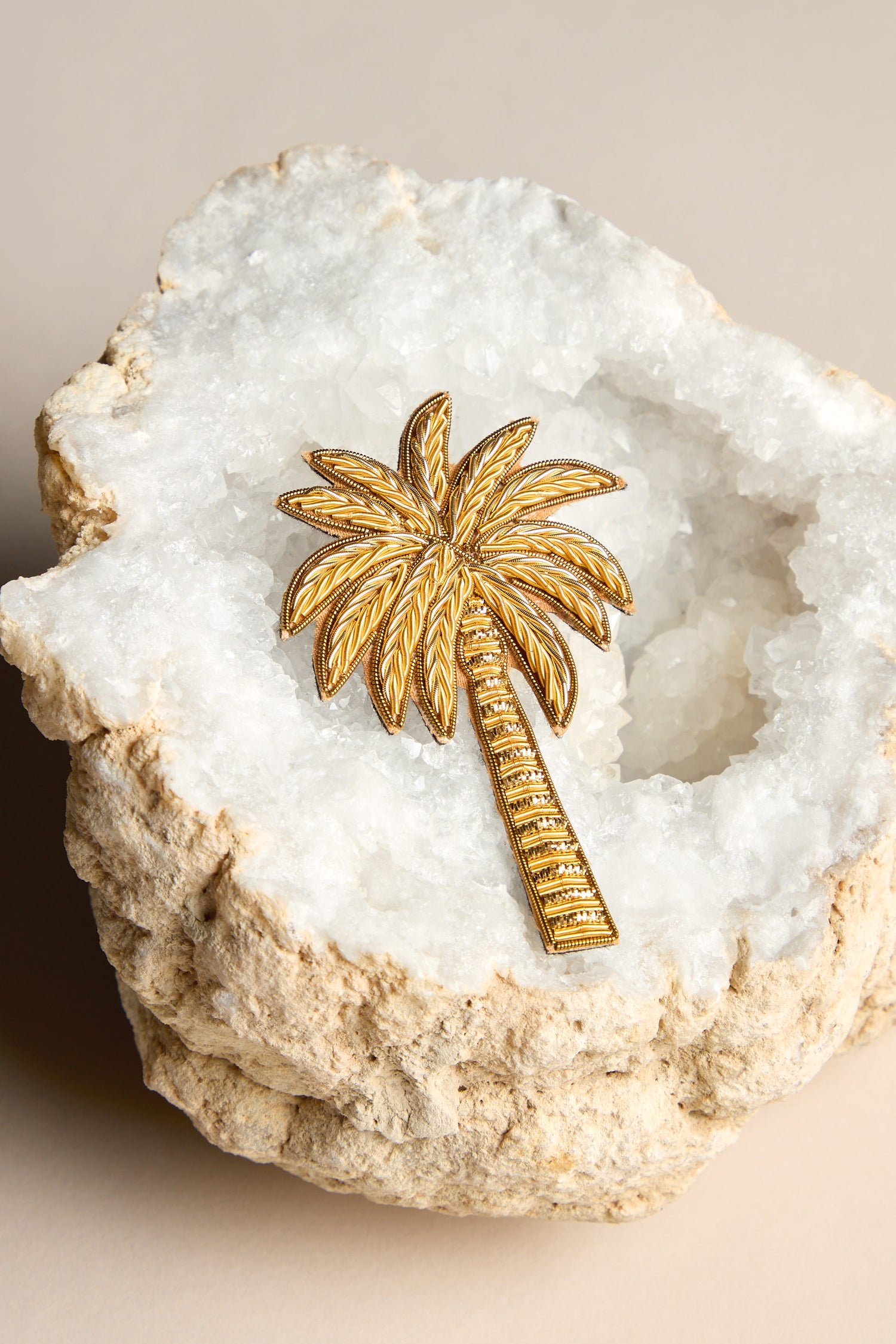 A glamorous Golden Palm Brooch rests on a rough, beige-edged white geode, evoking the allure of tropical palms.