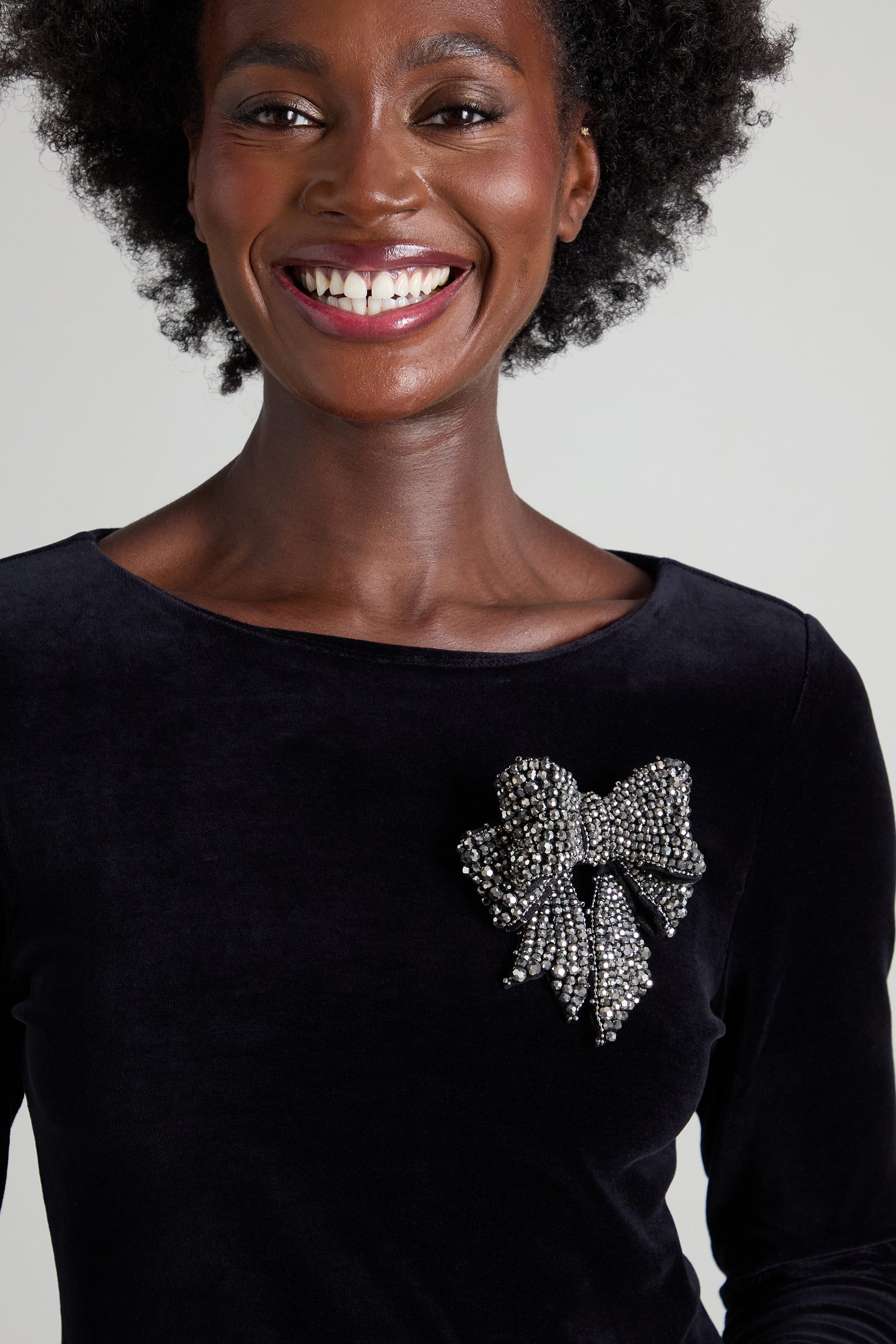 The person is smiling, adorned in a black top that exudes elegance, featuring the Bow Brooch as a striking large beaded embellishment.