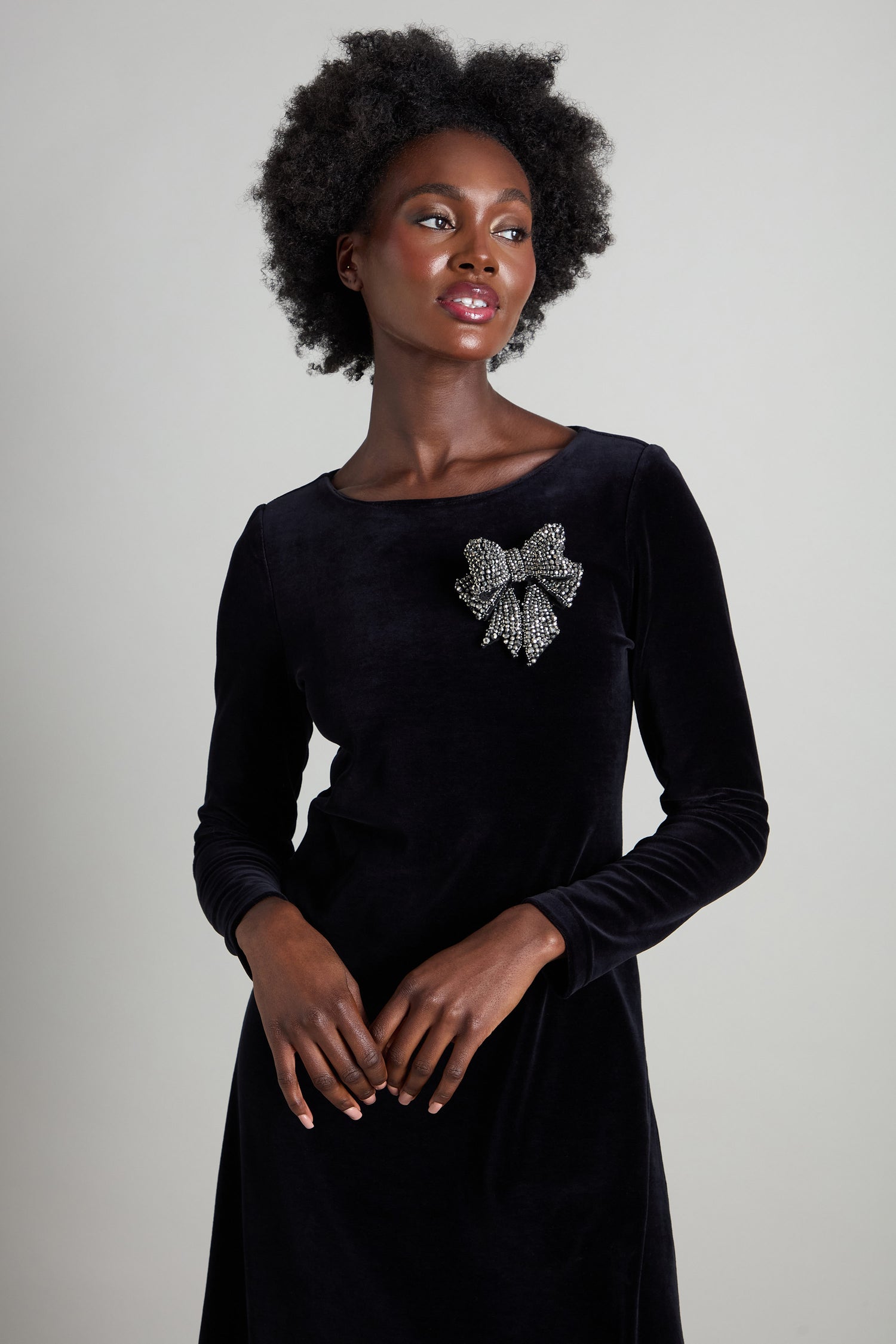 A person poses against a simple backdrop, radiating sophistication in a long-sleeved black dress accentuated by the Bow Brooch, a bold and eye-catching accessory on the chest. Their short curly hair complements their serene expression, enhancing the elegant ensemble.