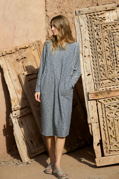 A woman wearing the Textured Stripe Jersey Dress and sandals stands outdoors beside carved wooden panels leaning against a textured wall.