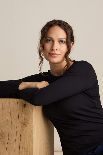 A woman with long, wavy brown hair, wearing the Viscose Jersey Relaxed Top – a true wardrobe staple – leans her arm on a wooden block and looks at the camera.