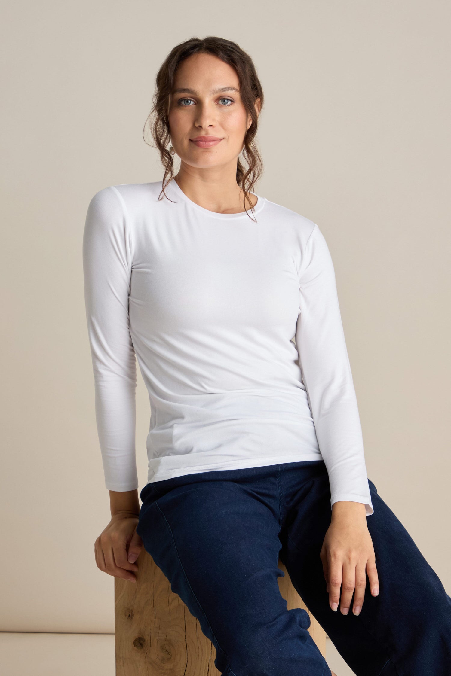 A person with brown hair wearing a white Viscose Jersey Relaxed Top and blue pants sits on a wooden block against a plain background.