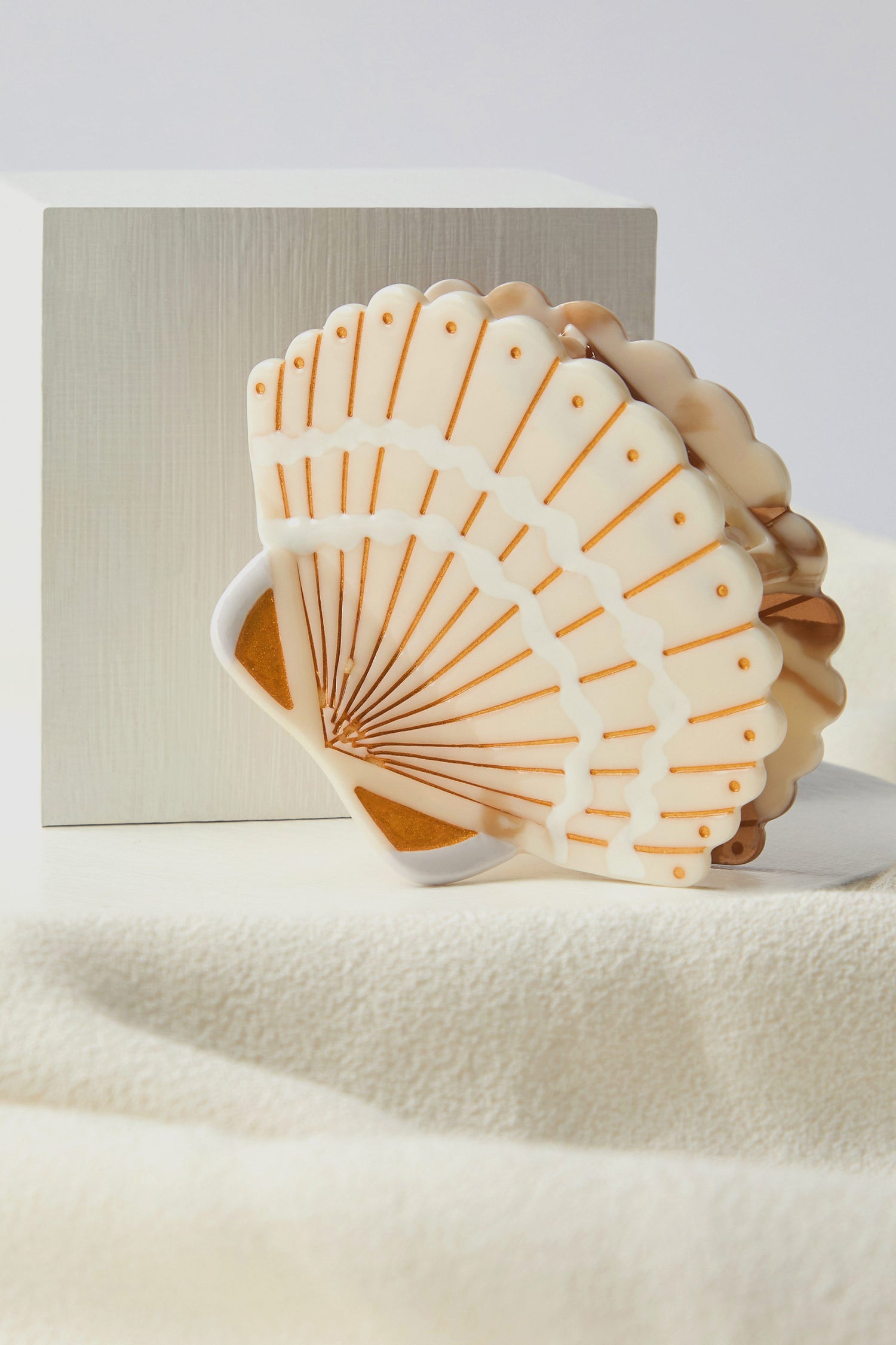 A white fan-shaped object, akin to a Hand Painted Seashell Hair Clip with orange accents, rests elegantly against a white cube on cream fabric. Its design suggests sustainable craftsmanship and serves as an eco-friendly accessory for the discerning eye.