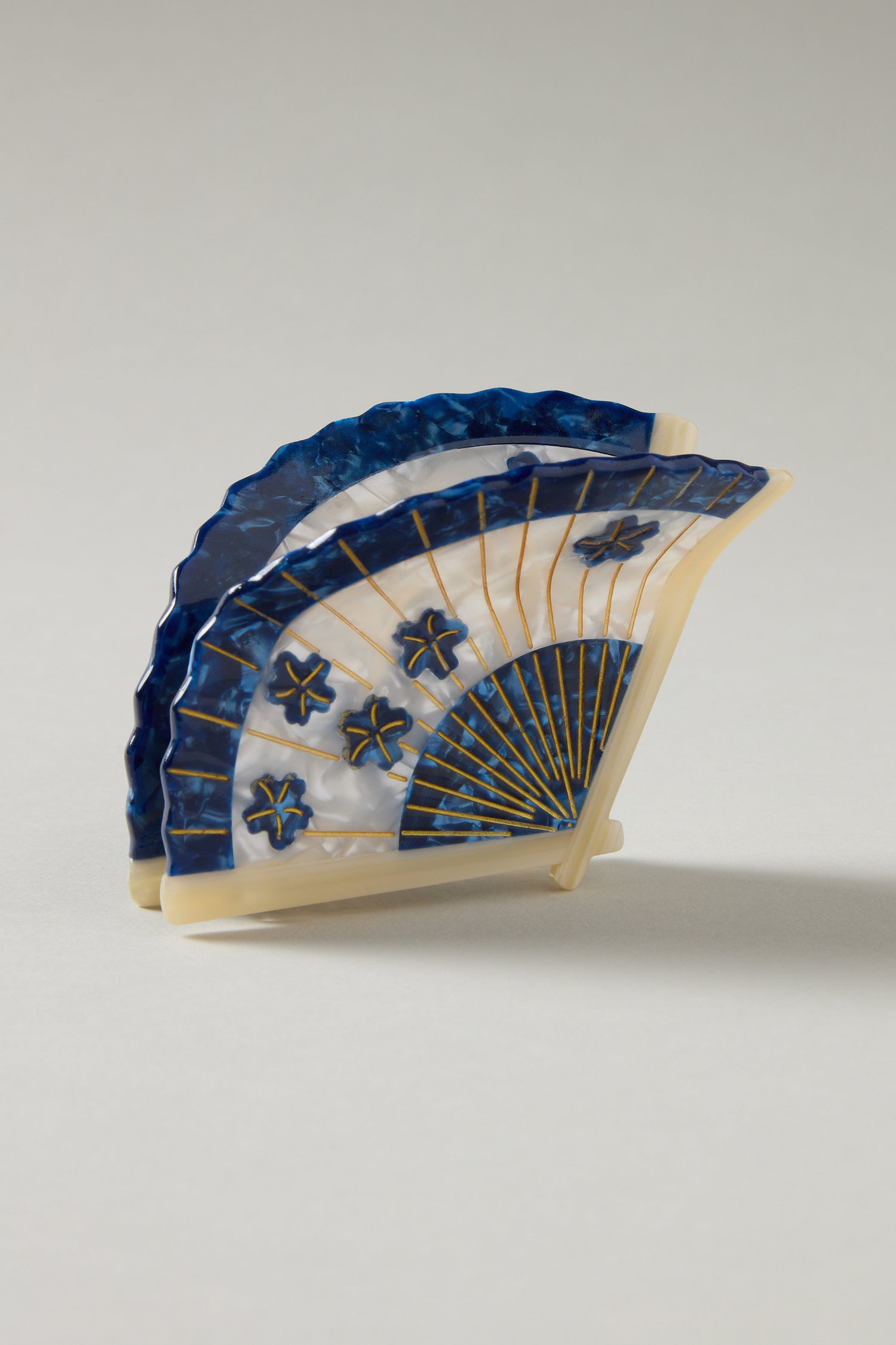 The Hand Painted Blossom Fan Hair Clip, made with eco-friendly techniques, features a blue and white pattern adorned with gold lines and star-shaped designs. It stands against a plain background, highlighting the elegance of sustainable artistry.