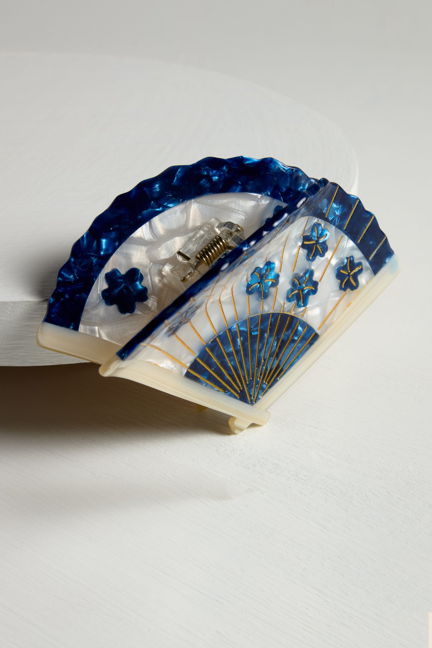 The Hand Painted Blossom Fan Hair Clip, made from biodegradable acetate, showcases blue and white floral and line designs. Its eco-friendly design adds a sustainable element while elegantly sitting on a white surface.