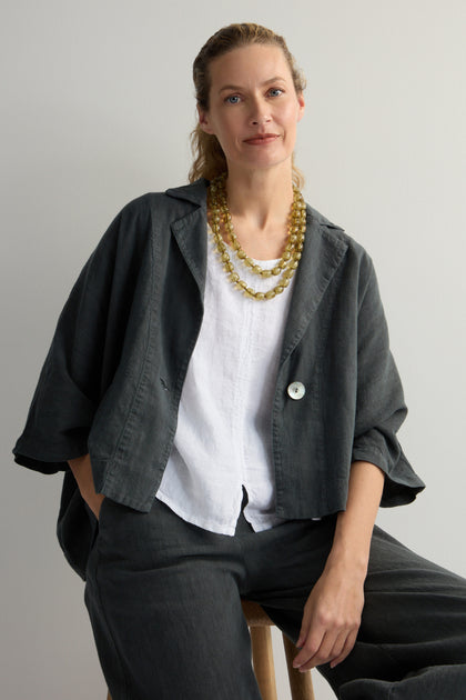 A person with long hair, in a dark oversized jacket and white shirt, sits serenely on a stool against a plain background. A Long Resin Bead Necklace lends elegance to the look, with versatile accessories subtly hinting at earthy khaki tones.