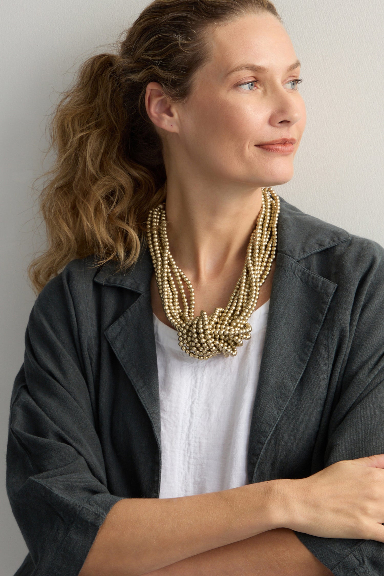 A woman with long hair wears a Gold Knotted Bead Necklace and a dark jacket over a white top, looking to her left.