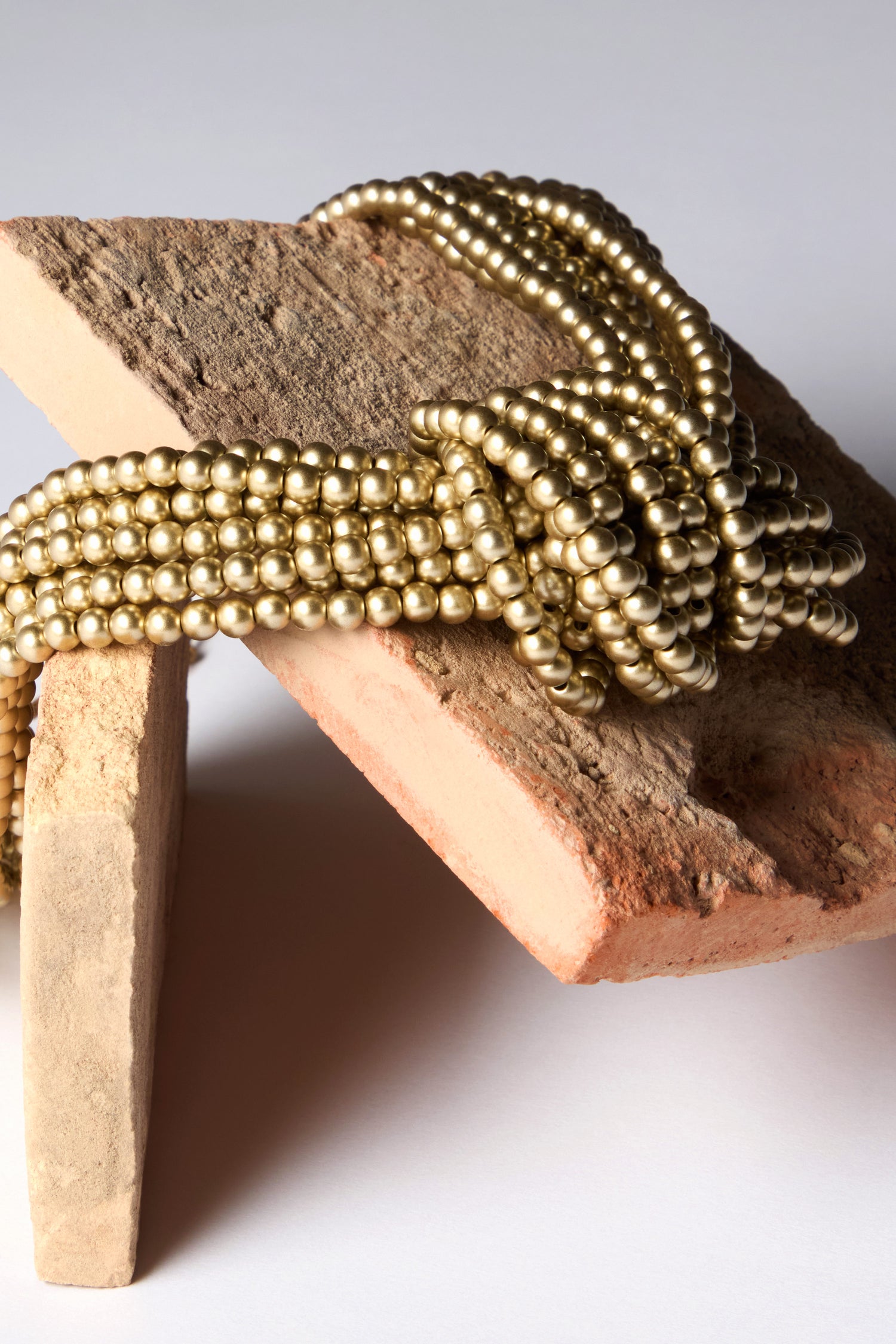 The Gold Knotted Bead Necklace with golden-toned beads gracefully drapes over two textured bricks against a plain backdrop.