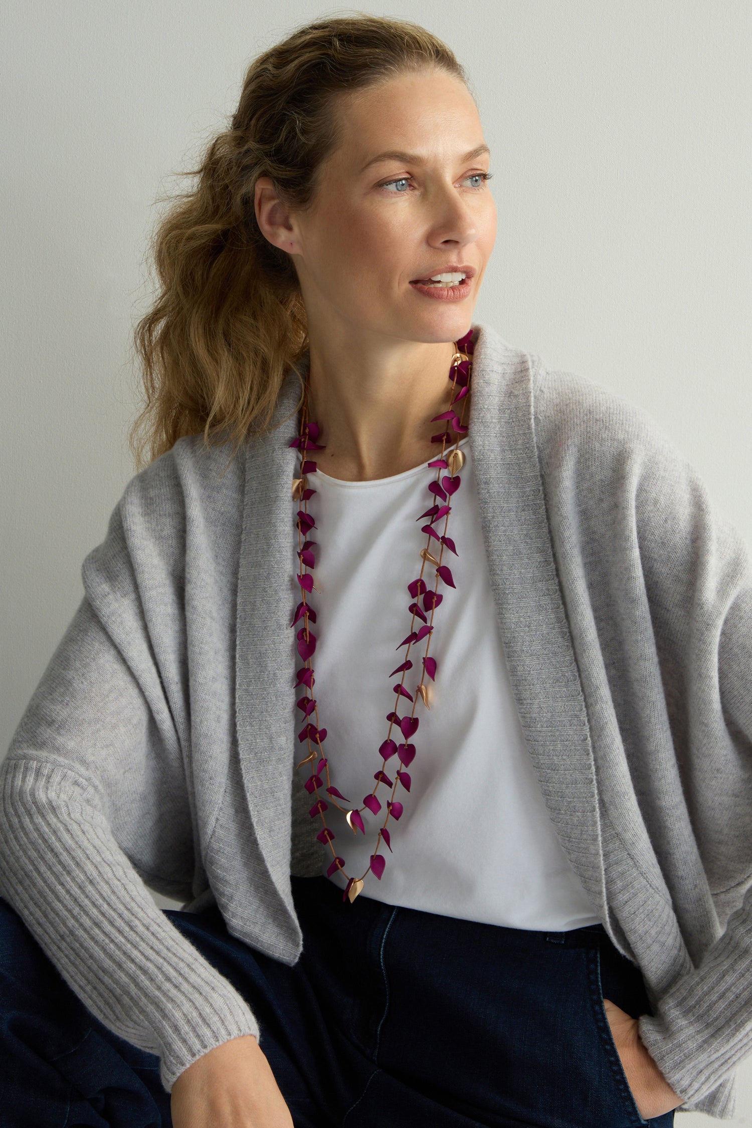 A blond-haired person is wearing a gray sweater, white shirt, and a Fuchsia Cascade Leaves Necklace with gold-toned details, sitting against a light background.