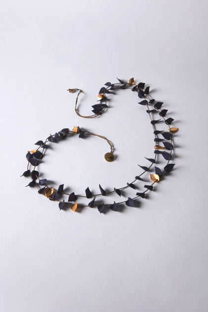 The Navy Cascade Leaves Necklace showcases alternating black and gold metal leaf-shaped elements, elegantly arranged on a light gray surface.