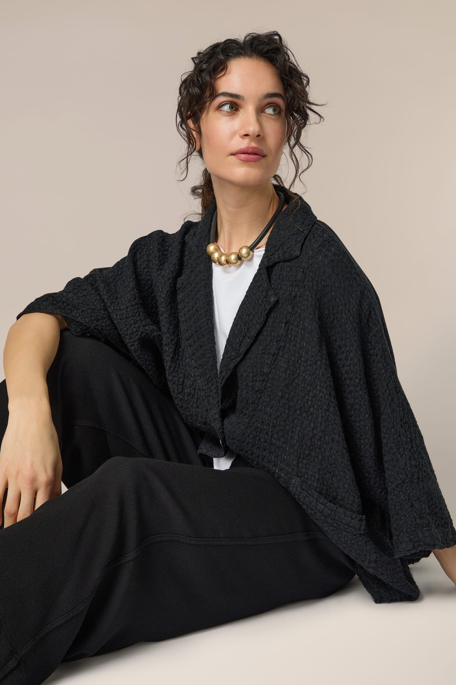 A woman is sitting on the floor wearing black pants and a black sweater, accessorized with a Hammered Gold Spheres Necklace.