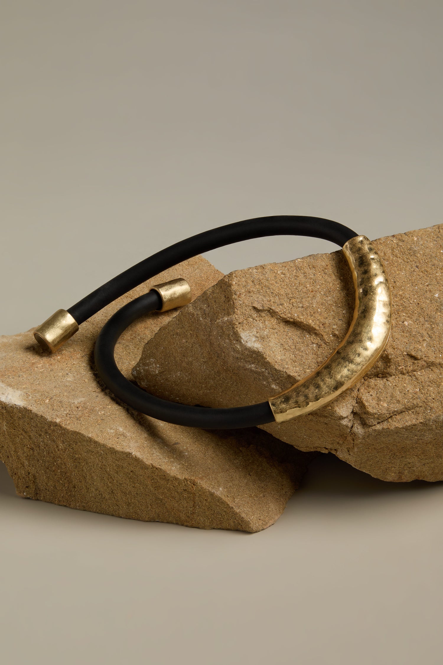 The Hammered Necklace, featuring a contemporary black and gold design with a hammered tubular pendant, is elegantly showcased on two rough, brown stone pieces.