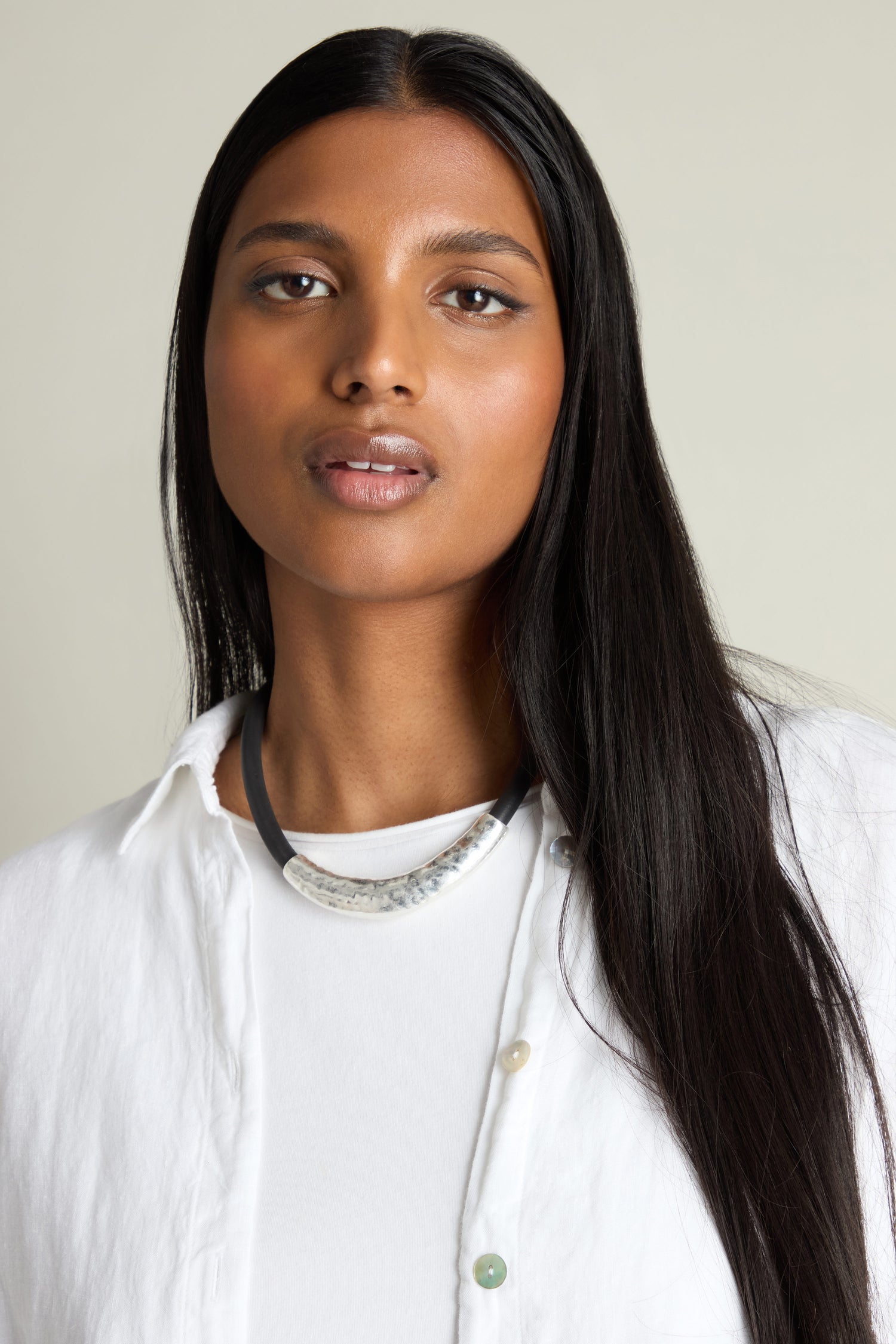 A person with long black hair, dressed in a white top and showcasing the Hammered Necklace, which features a striking metallic hammered pendant, set against a plain background.