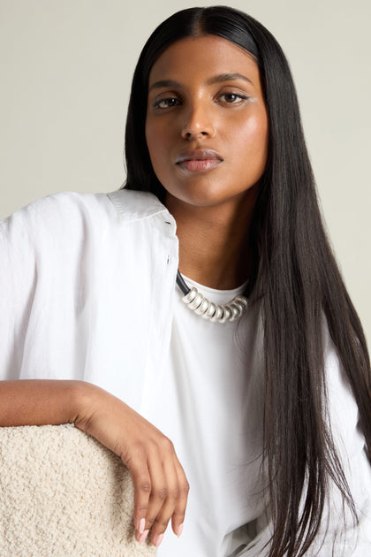 A person with long dark hair is wearing a white shirt and the Hammered Silver Cylinders Necklace, sitting against a neutral background. The eye-catching necklace adds a stylish touch to their look.