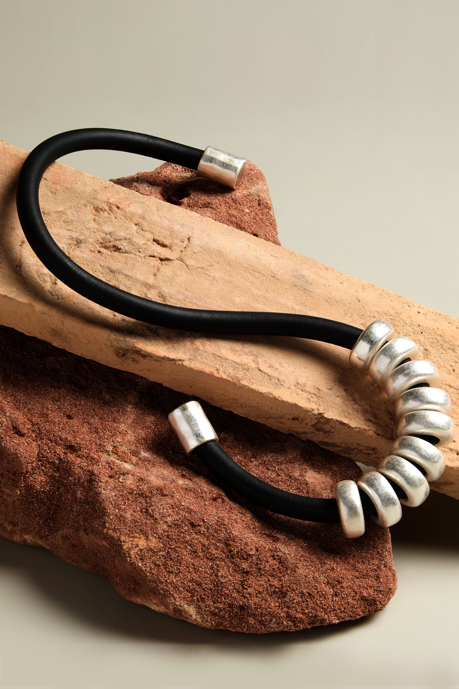 The Hammered Silver Cylinders Necklace, featuring brushed silver beads and a sleek black cable, is elegantly displayed on brown and beige rocks against a neutral background, accentuated by a subtle silver magnet closure.