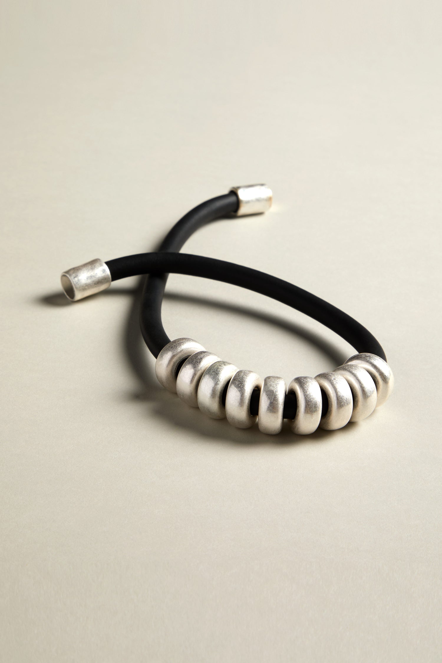 The Hammered Silver Cylinders Necklace offers a contemporary design with its black cord, adorned with multiple brushed silver loops and cylindrical accents. Its sleek look is enhanced by a stylish silver magnetic closure, set against a light gray background.