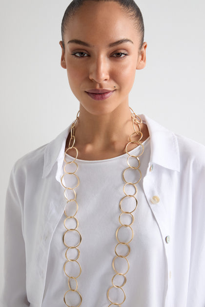 A person in a white shirt, smiling slightly against a plain background, wears the Long Metal Hoop Necklace—a bold statement piece resembling a gold circular-link design.