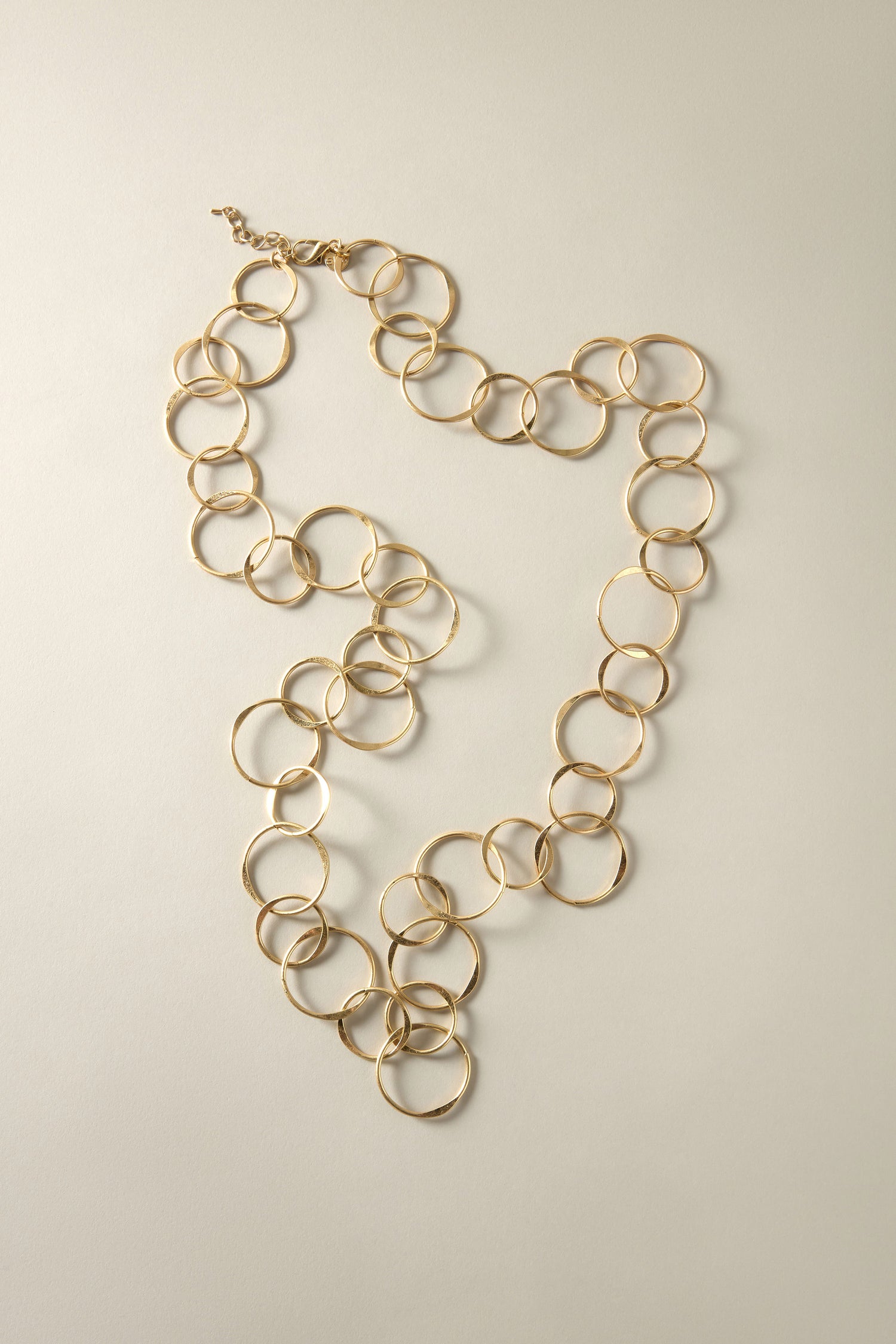 The Long Metal Hoop Necklace, featuring large circular links in a heart shape on a beige background, redefines elegance and garners admiration with its bold design.