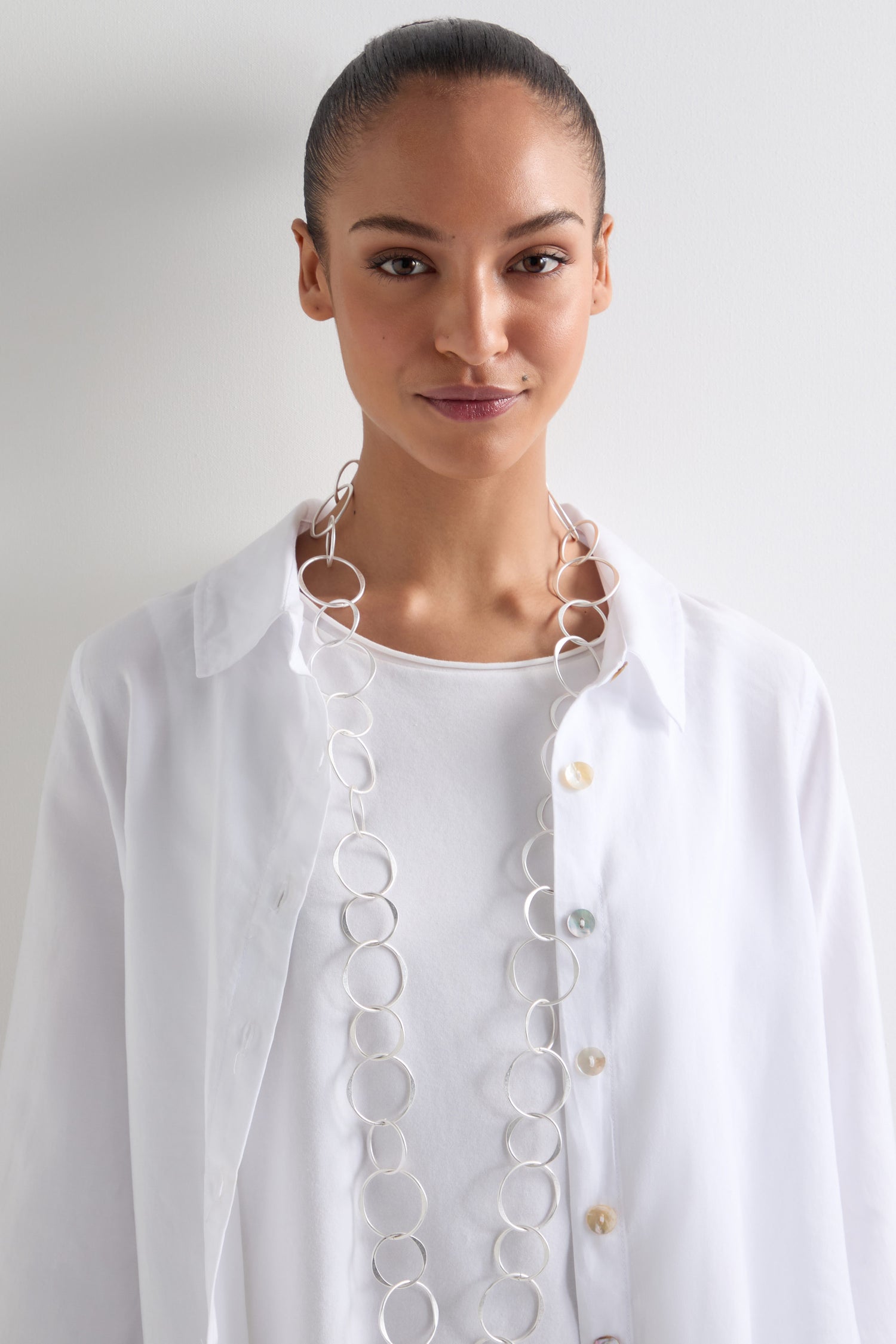 A person with their hair tied back wears a white shirt and a standout accessory—a Long Metal Hoop Necklace—set against a plain light background.