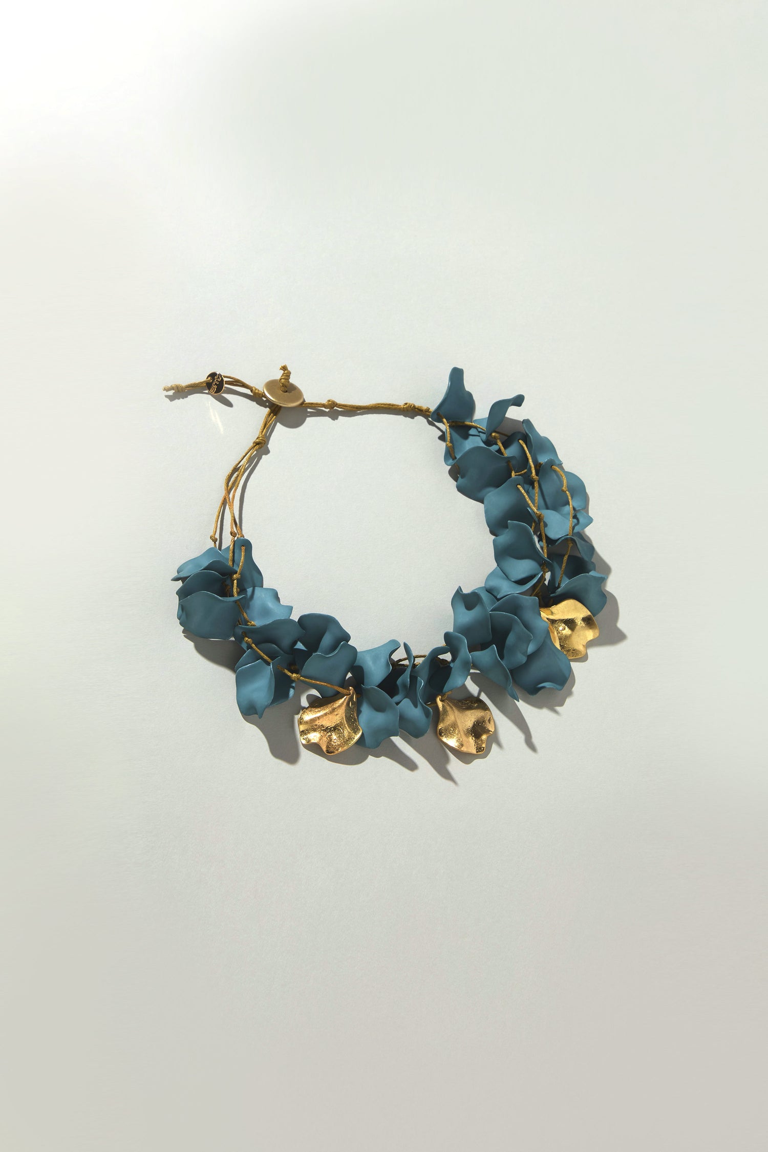 The Short Cascade Petal Necklace features blue petal-like shapes with gold accents. Its adjustable cord ensures a perfect fit against a light background, exuding organic elegance and serving as a timeless accessory.